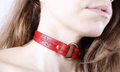 A close-up of a woman's neck and chin wearing a red choker necklace with a gold-colored metal ring.