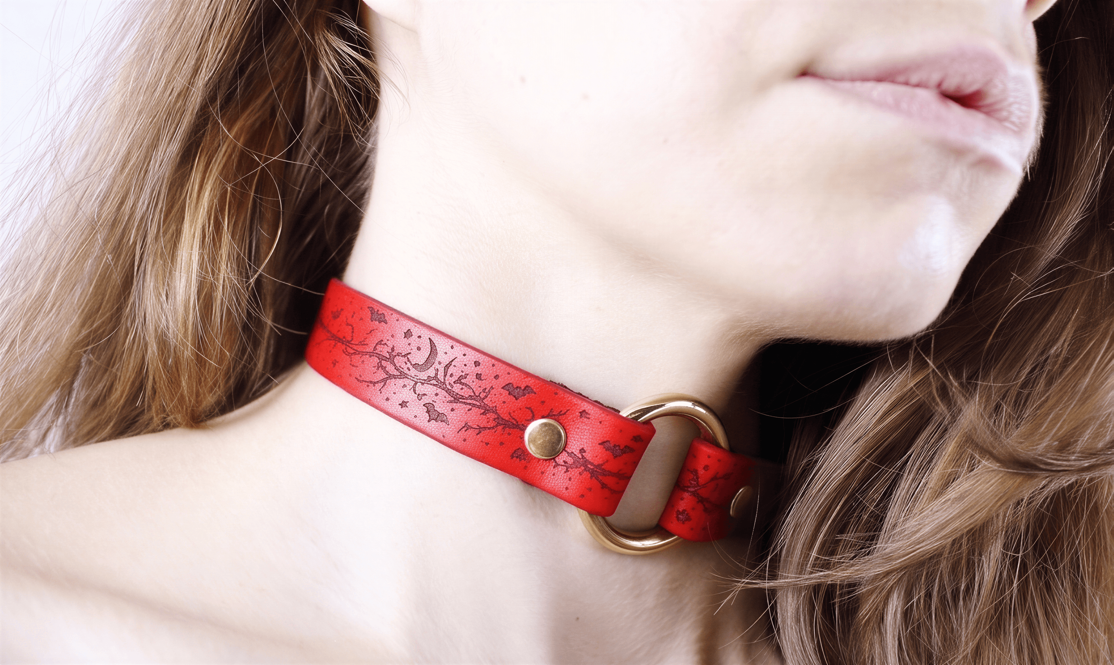 A close-up of a woman's neck and chin wearing a red choker necklace with a gold-colored metal ring.
