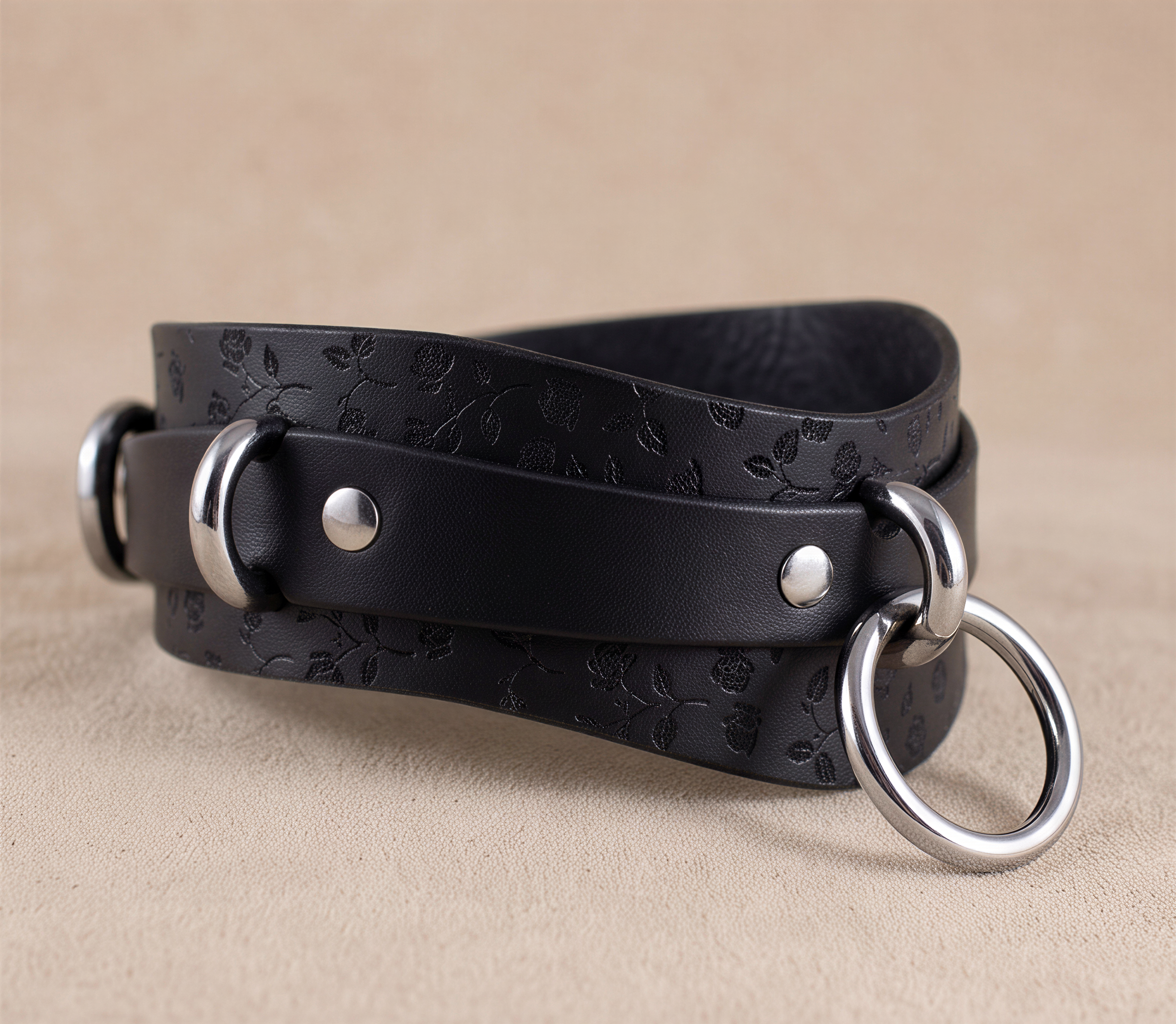 A black leather strap with metal rings and a floral pattern, resting on a beige surface.