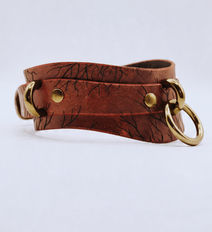 A brown leather strap with a metal ring and two brass buckles, featuring a distinctive tree-like pattern.