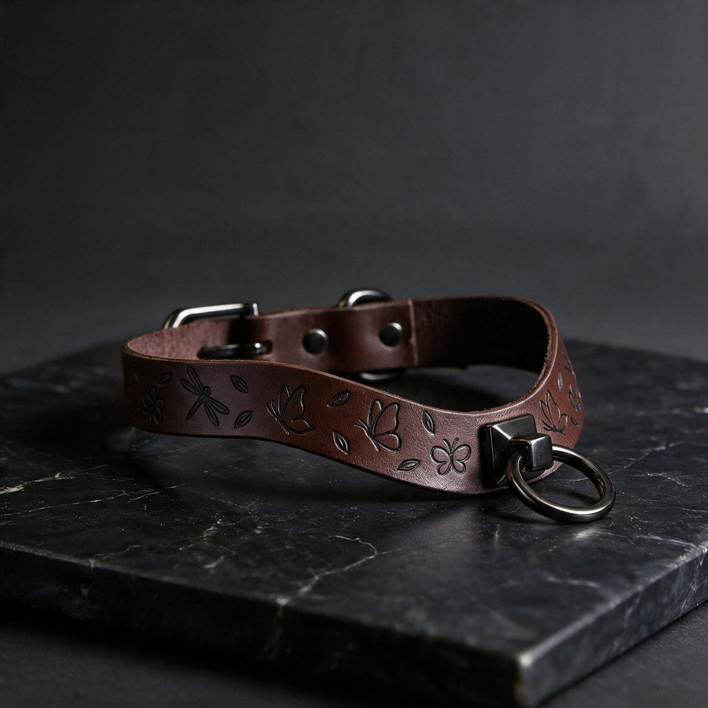 A brown leather collar with a metal ring and leaf designs on it, resting on a dark surface.