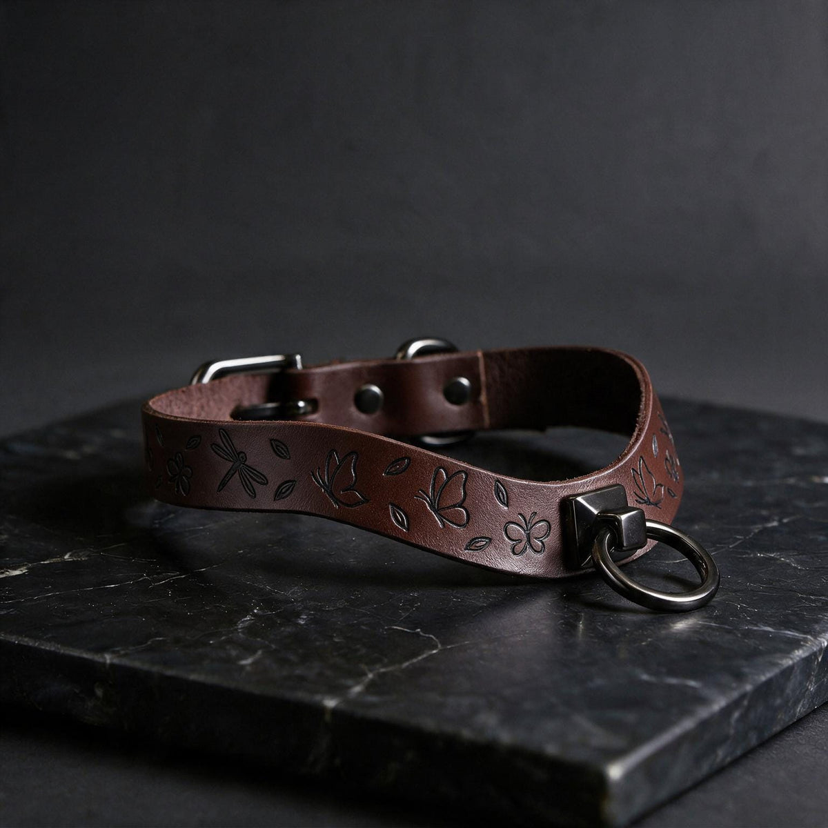 A brown leather collar with a metal ring and leaf designs on it, resting on a dark surface.