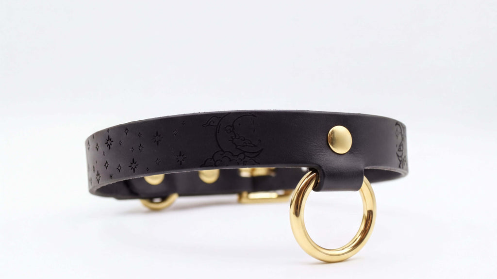 A black leather collar with a gold ring and a small moon-like design on the strap.