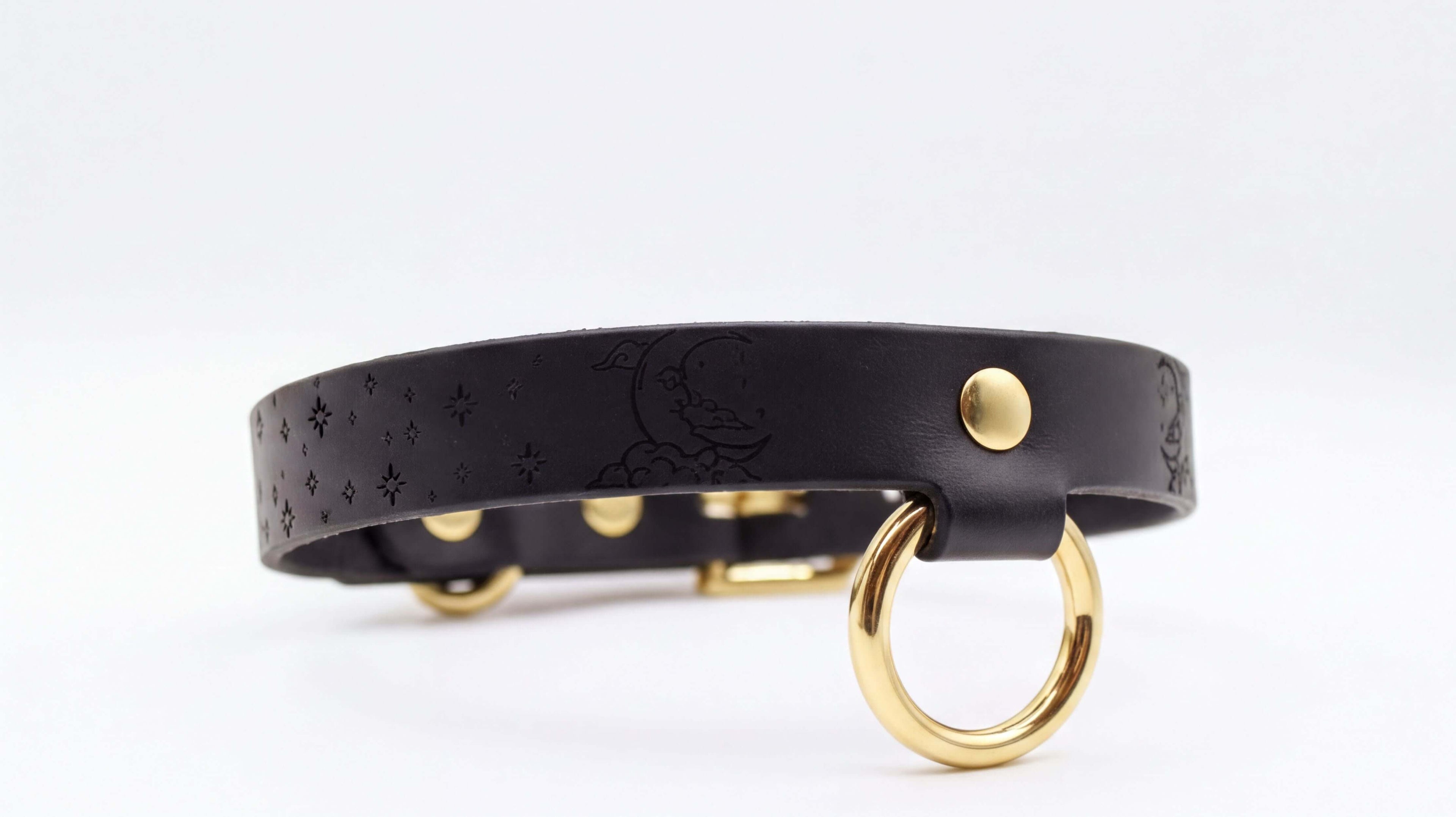 A black leather collar with a gold ring and a small moon-like design on the strap.