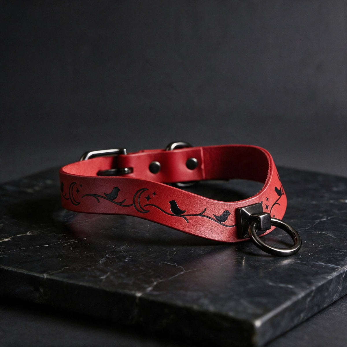 A red leather collar with a black bird design and a metal ring is displayed on a black marble surface.
