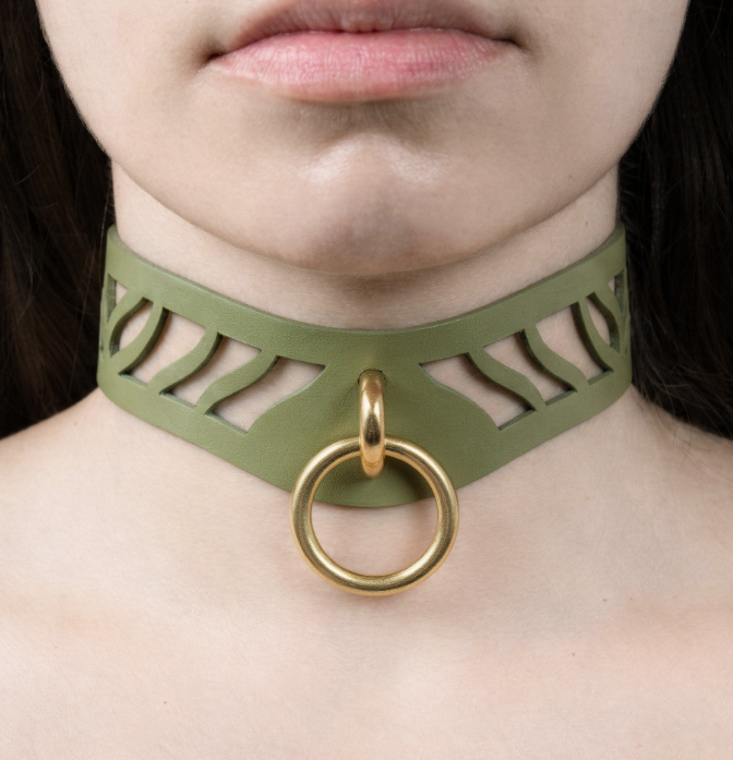 A close-up of a person's neck and chest area, featuring a green choker necklace with a gold ring and a cutout design.