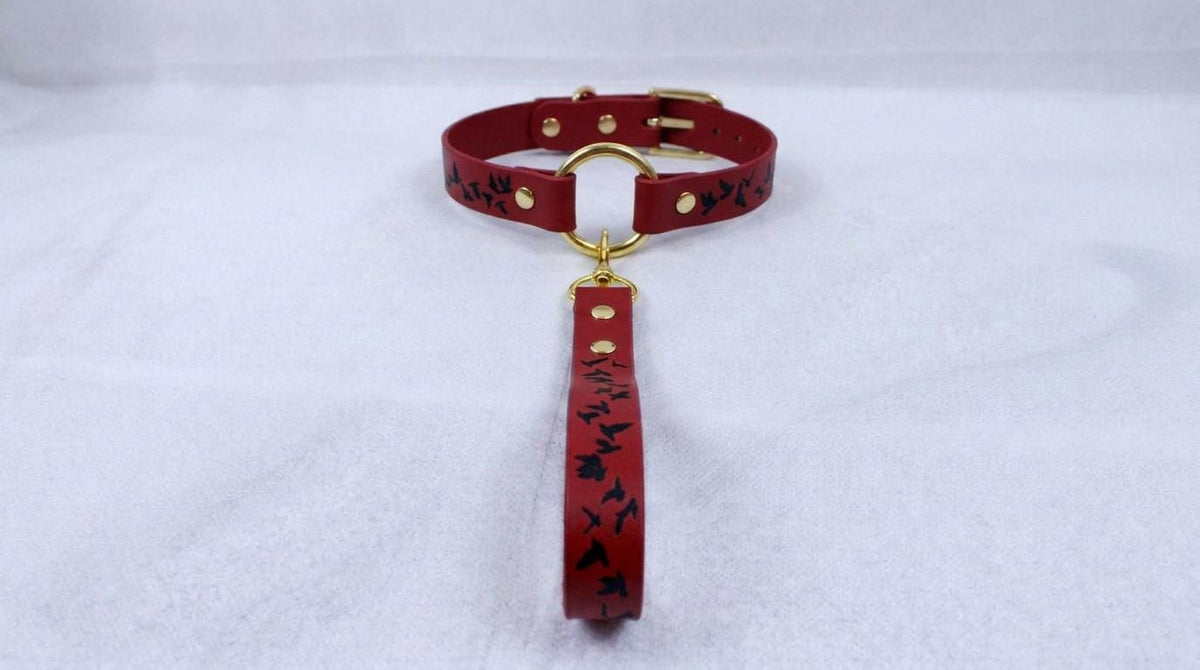 A red leather collar with a gold buckle and a black bird design on it, resting on a white surface.