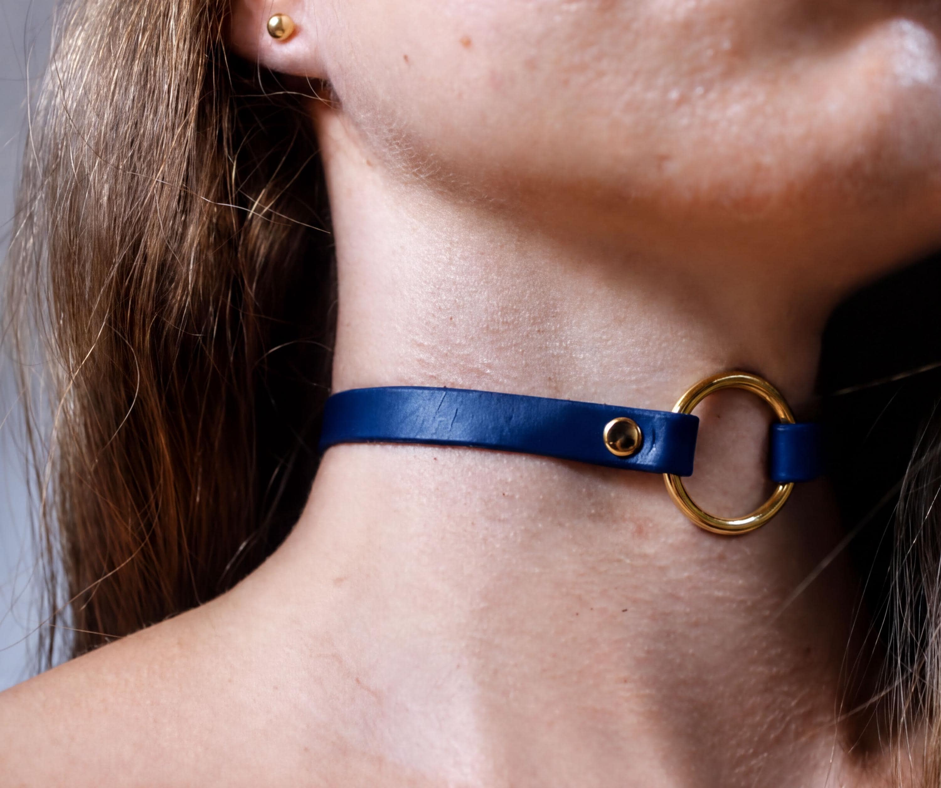 A close-up view of a person's neck and chin area, featuring a blue leather choker necklace with a gold-colored circular clasp.