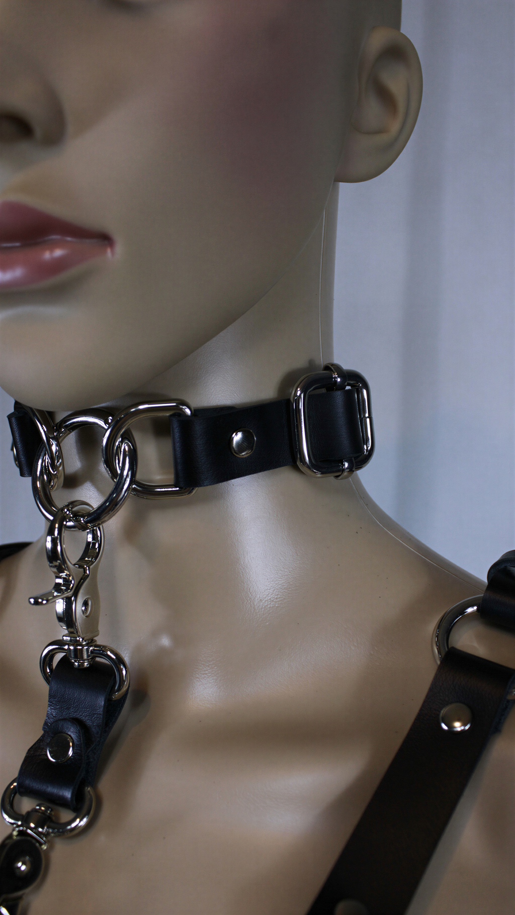 A close-up view of a mannequin wearing a black leather choker necklace with metal rings and buckles.