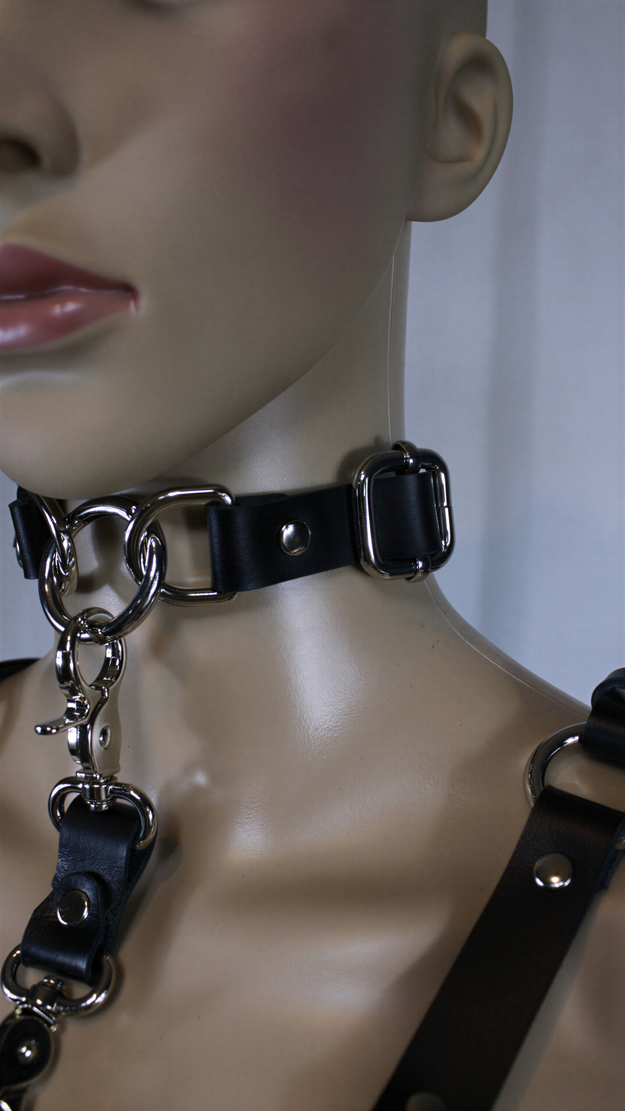 A close-up view of a mannequin wearing a black leather choker necklace with metal rings and buckles.