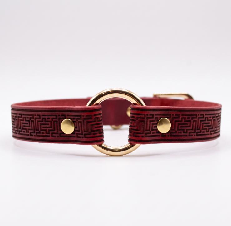 A burgundy leather collar with a gold-colored metal ring and two gold-colored metal fasteners.