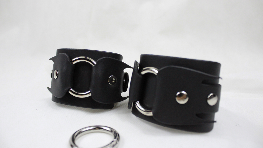 A pair of black leather cuffs with metal rings and buckles, placed on a white surface.