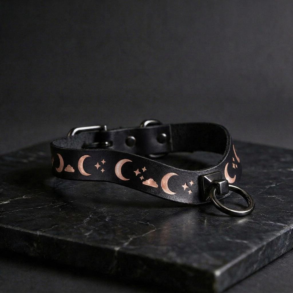 A black leather collar with a moon and star pattern design, featuring a metal ring with a clasp.