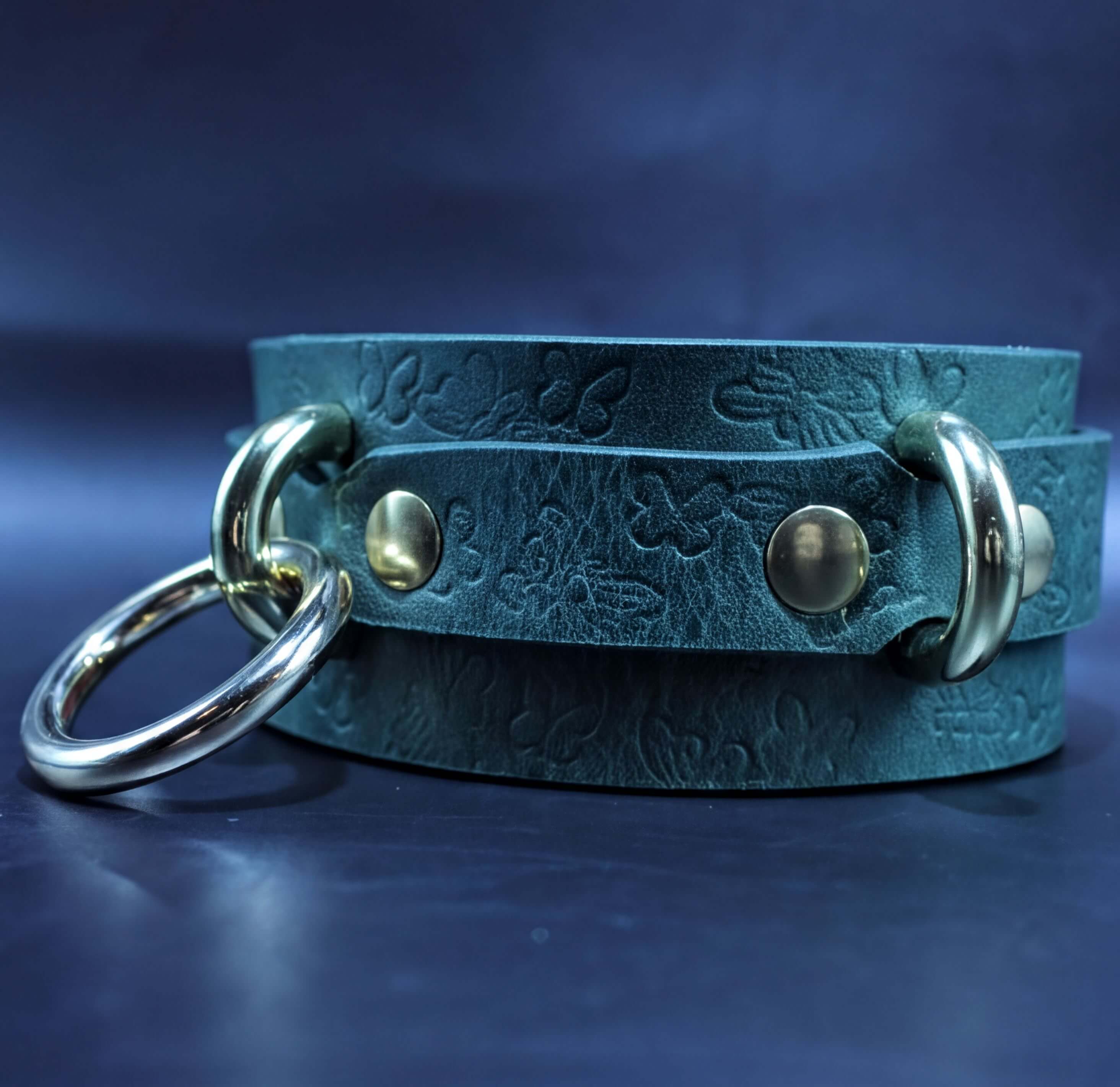 A close-up image of a green leather choker necklace with a metal ring and two gold-colored buckles.