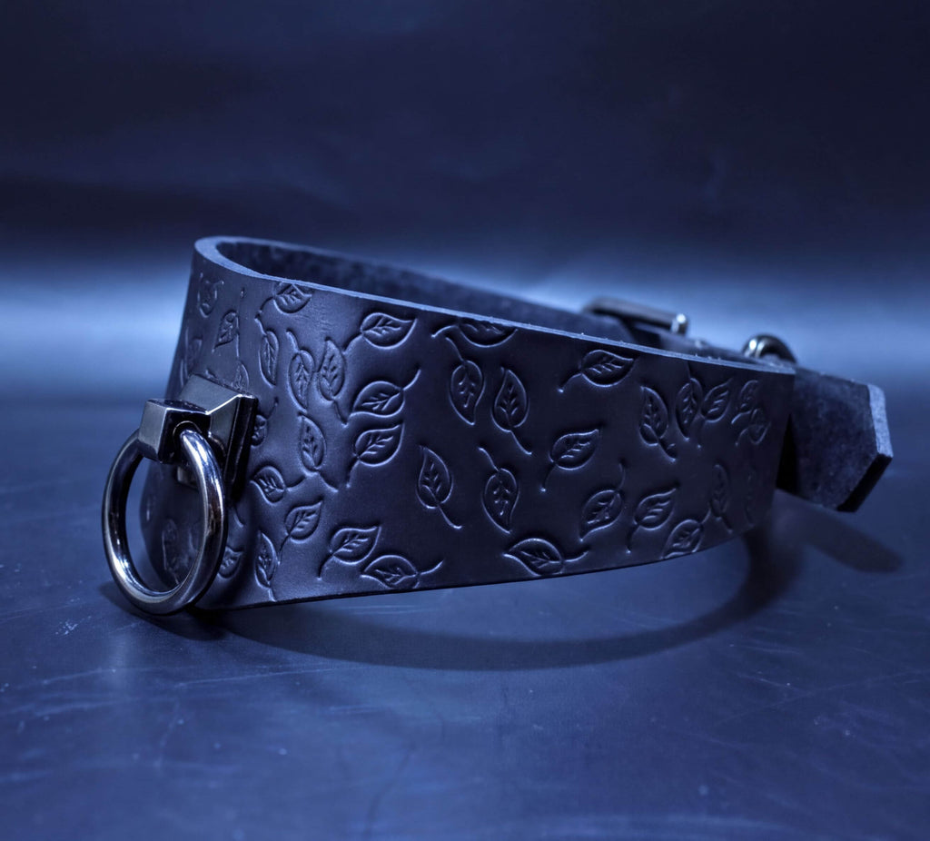 A black leather choker with a leaf pattern embossed on it, featuring a metal ring and buckle.