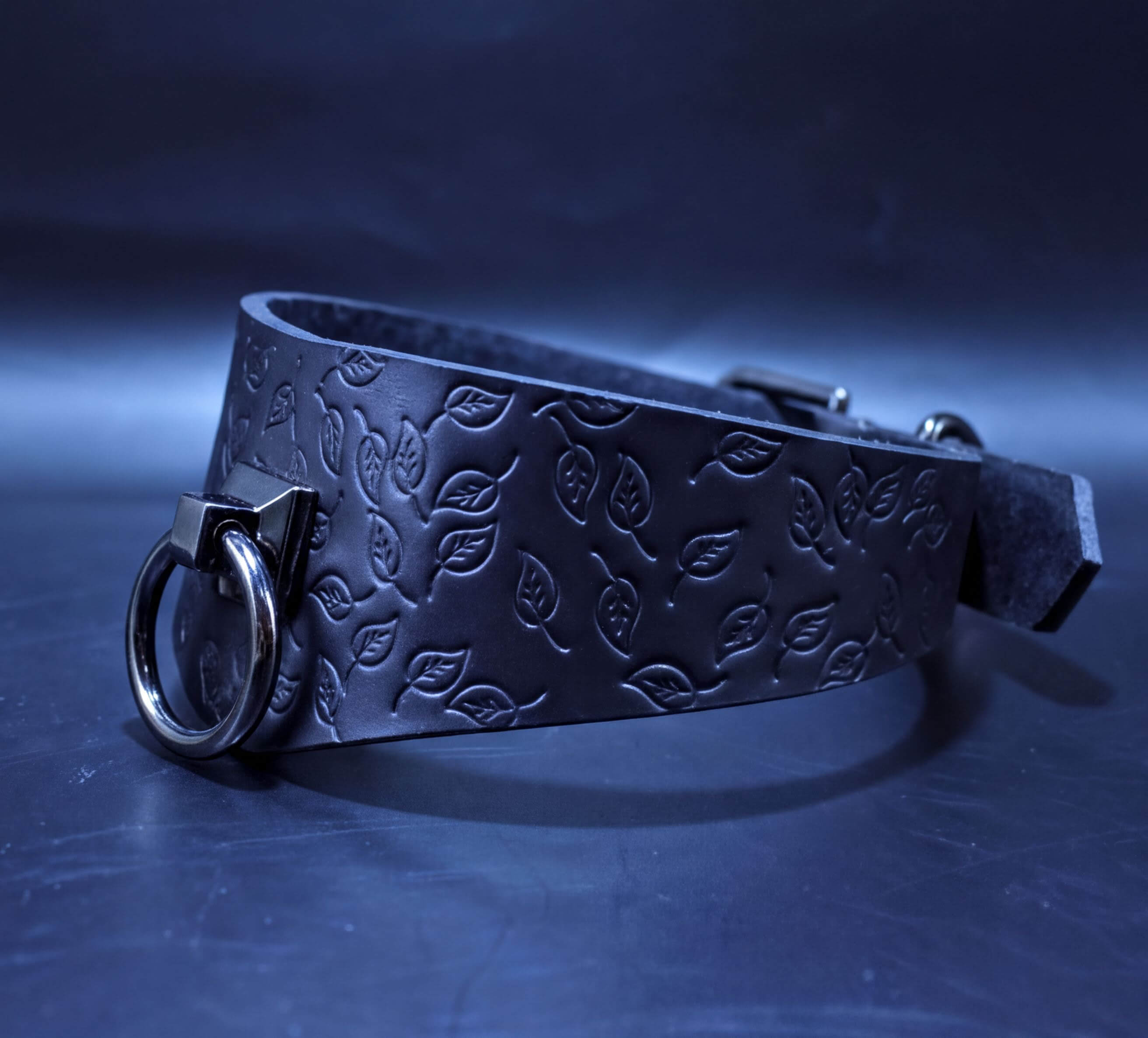 A black leather choker with a leaf pattern embossed on it, featuring a metal ring and buckle.