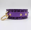 A purple leather collar with a floral pattern and gold-colored metal rings.