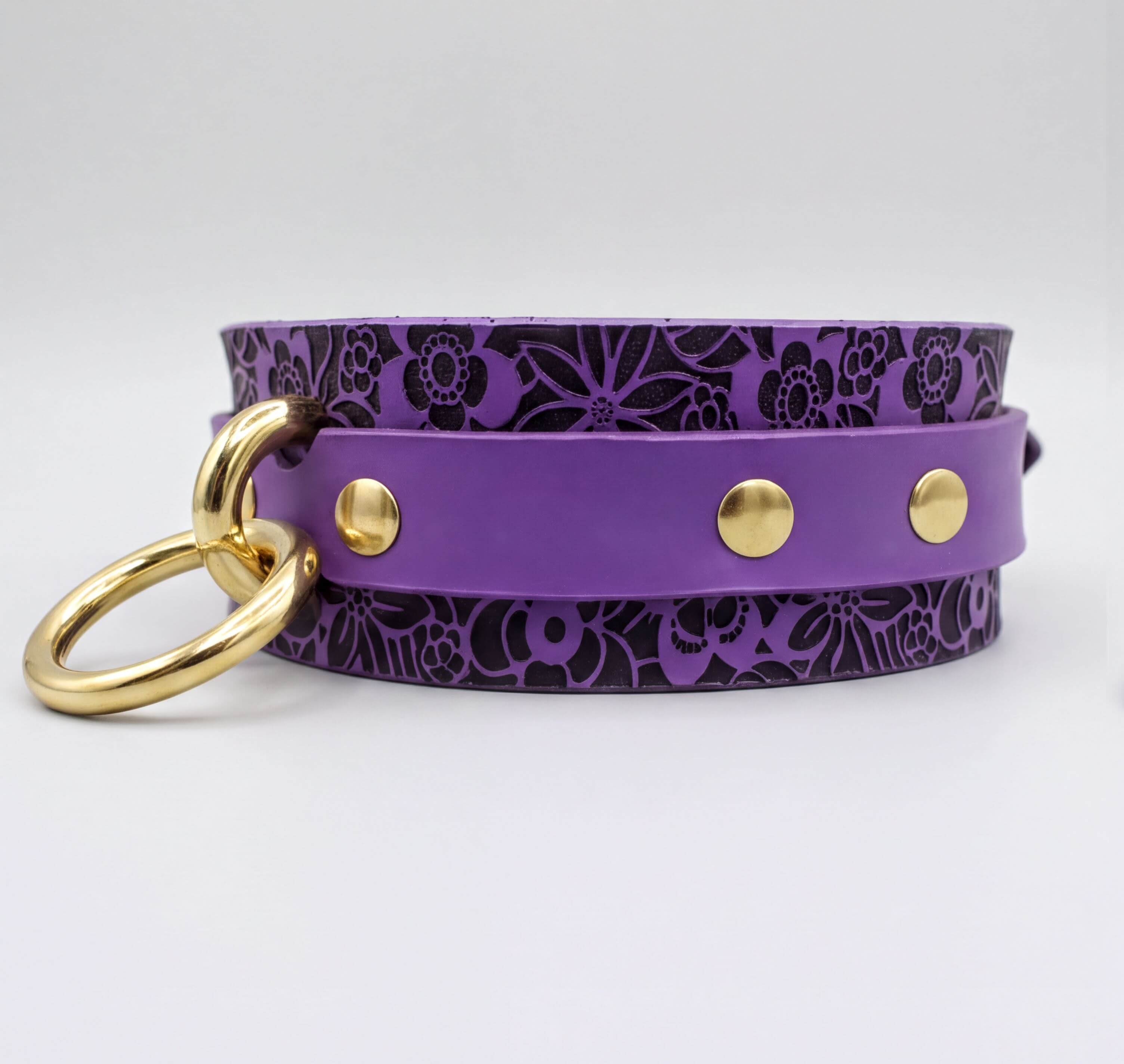 A purple leather collar with a floral pattern and gold-colored metal rings.