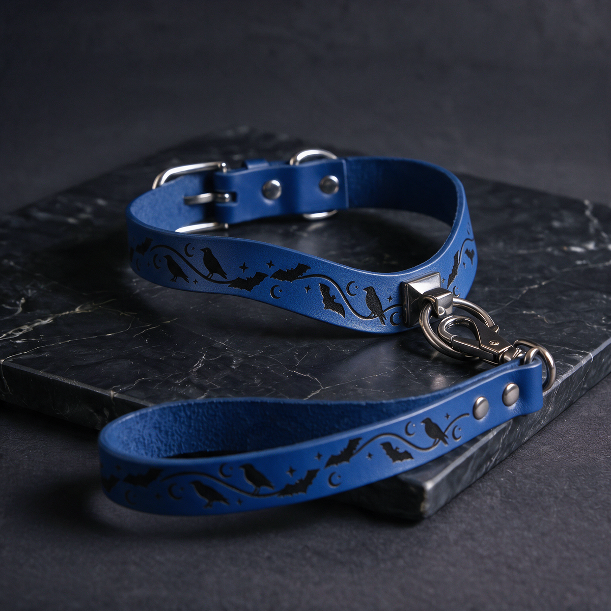 A blue leather dog collar with a bat and moon design, featuring a silver buckle and a black handle, is displayed on a dark surface.