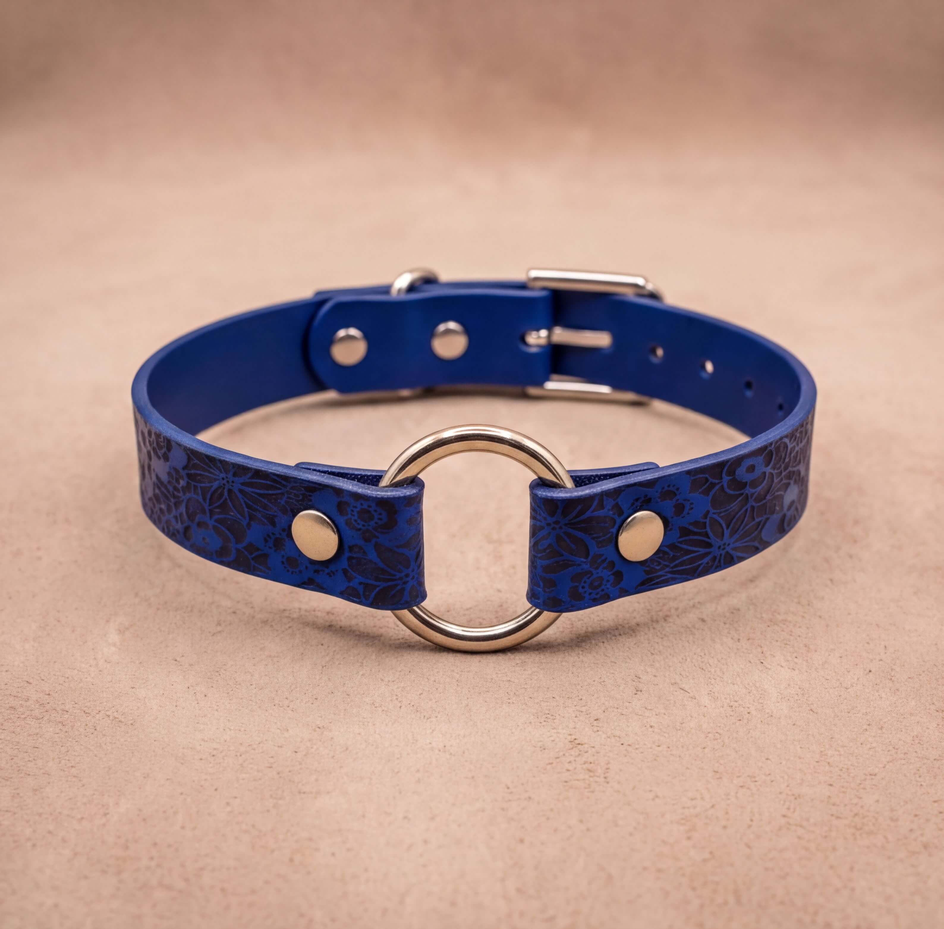 A blue leather collar with a metal ring and buckle, placed on a beige background.