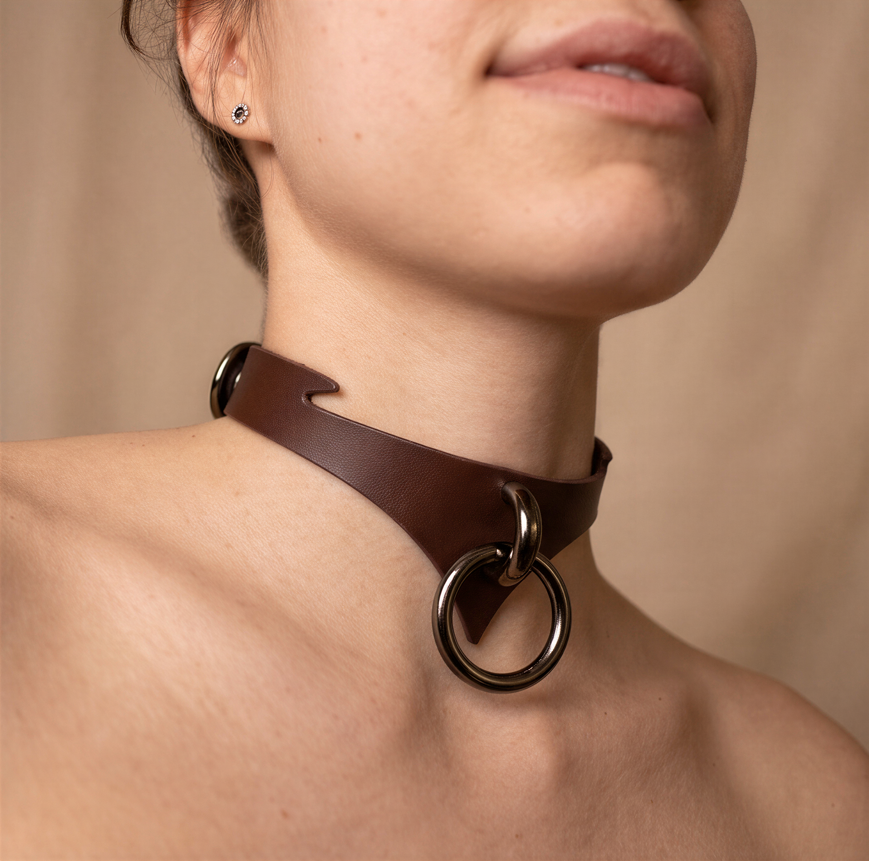 A close-up of a woman's neck and chest area wearing a brown leather choker necklace with a metal ring.