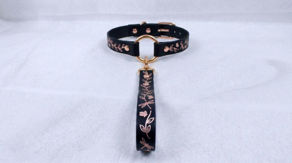 A black collar with a gold-colored metal clasp and a floral pattern on the strap is displayed against a white background.