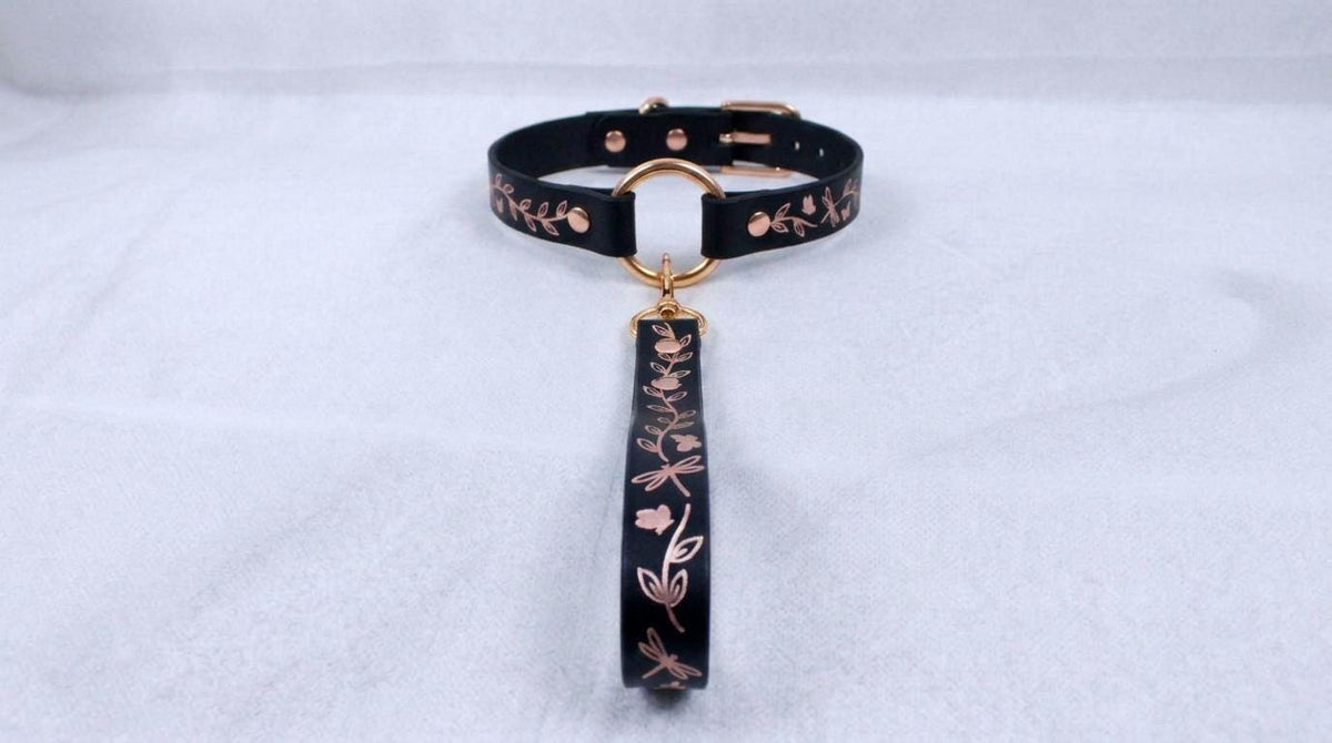 A black collar with a gold-colored metal clasp and a floral pattern on the strap is displayed against a white background.
