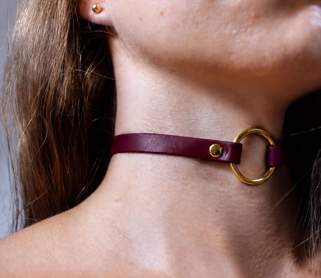A close-up view of a person's neck and chin area, featuring a burgundy leather choker necklace with a gold ring.