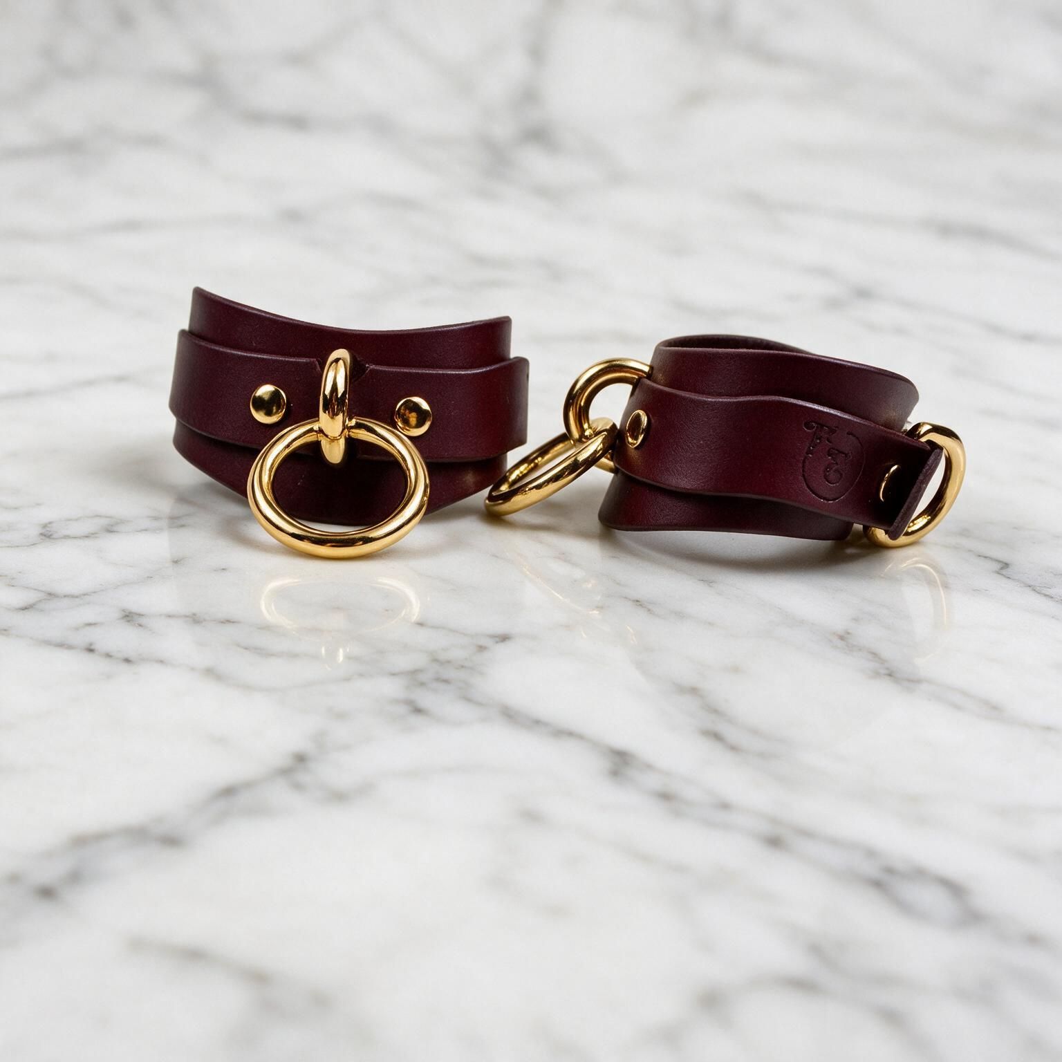 A pair of burgundy leather cinch straps with gold metal rings, placed on a marble surface.