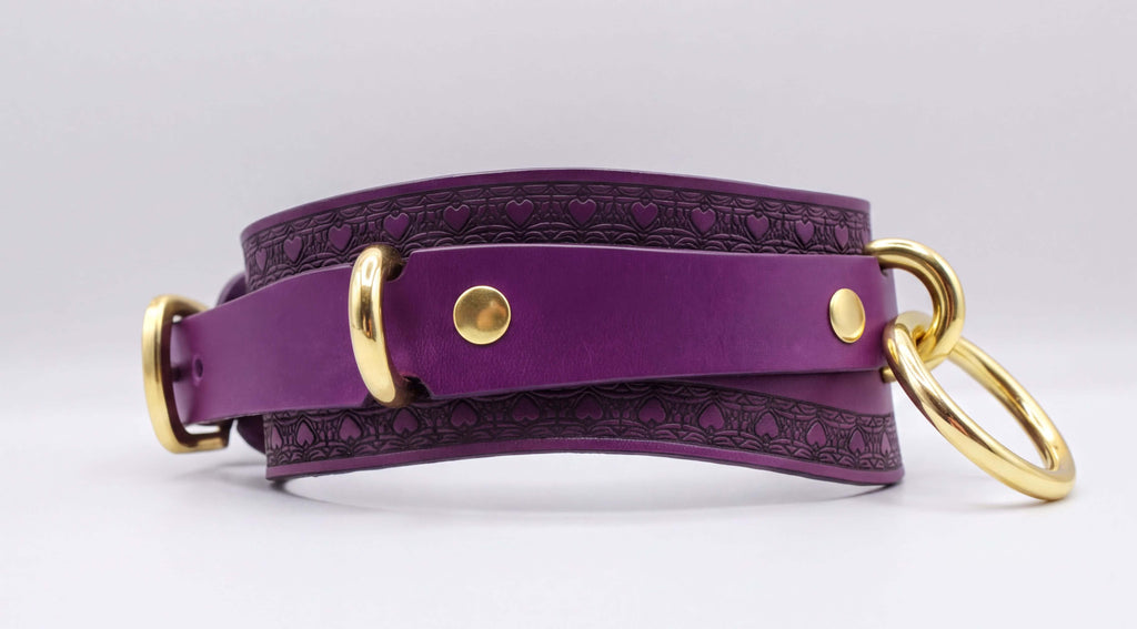 A purple leather choker collar with gold hardware, including a buckle and two rings, is shown in the image.