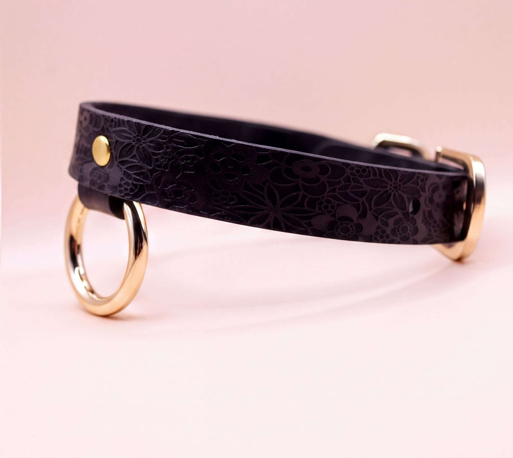 A black leather collar with a gold-colored metal ring on the front, set against a pink background.