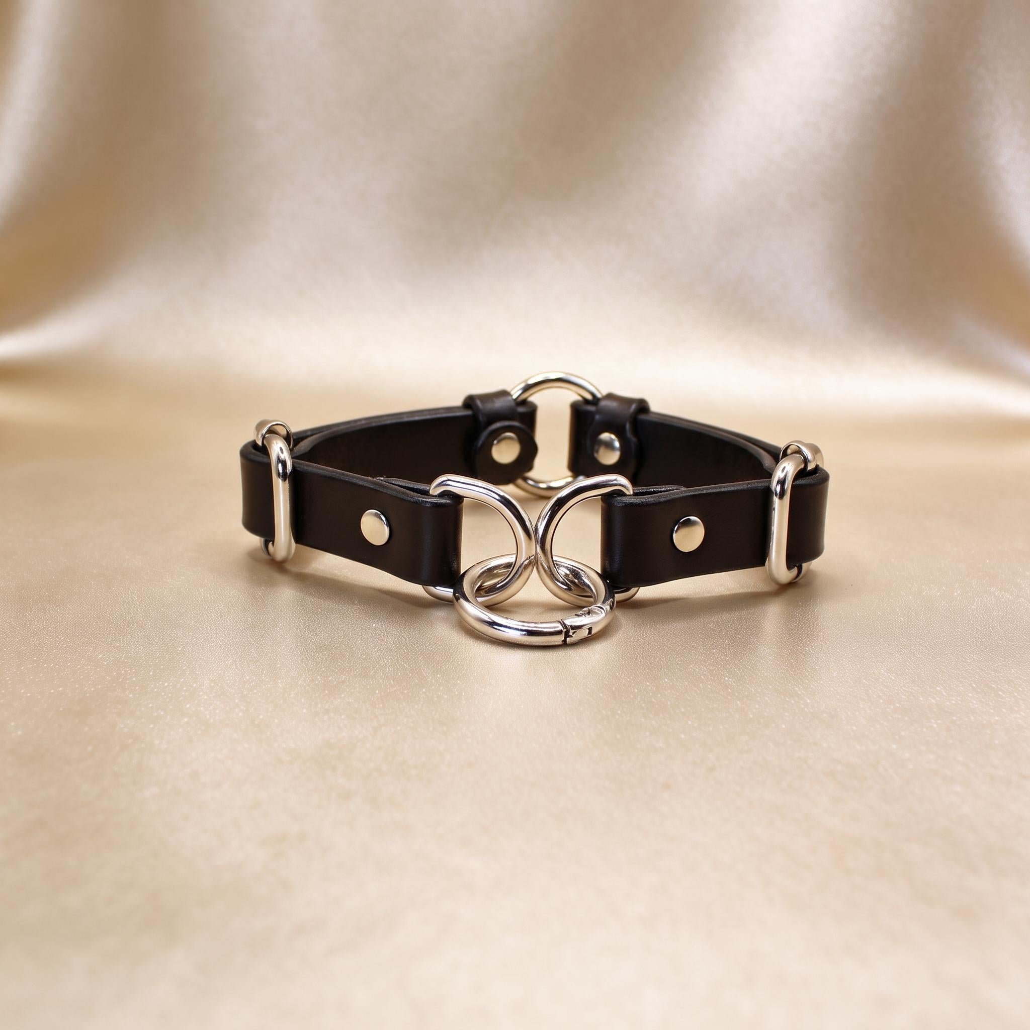 A black leather choker necklace with silver metal rings and buckles, displayed on a beige background.