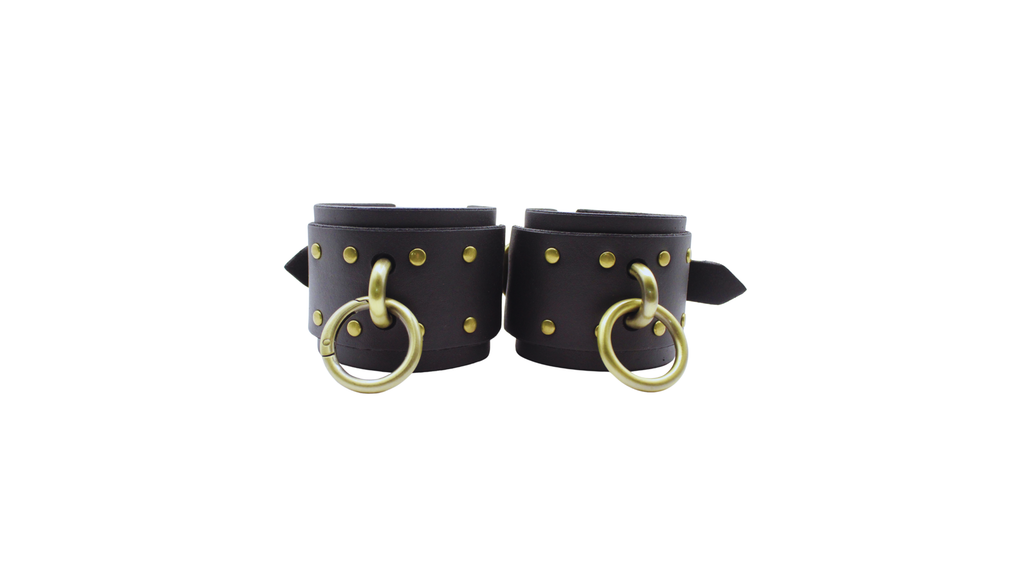 A pair of black leather wrist cuffs with gold-colored metal rings and studs.