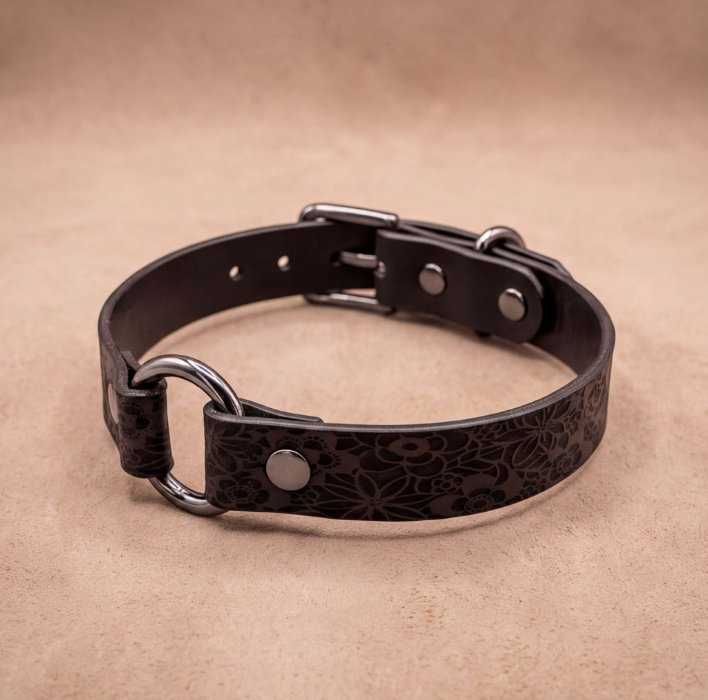 A black leather collar with metal rings and buckles, resting on a beige background.