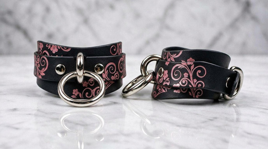 a pair of black leather straps with floral designs and metal rings, placed on a marble surface.