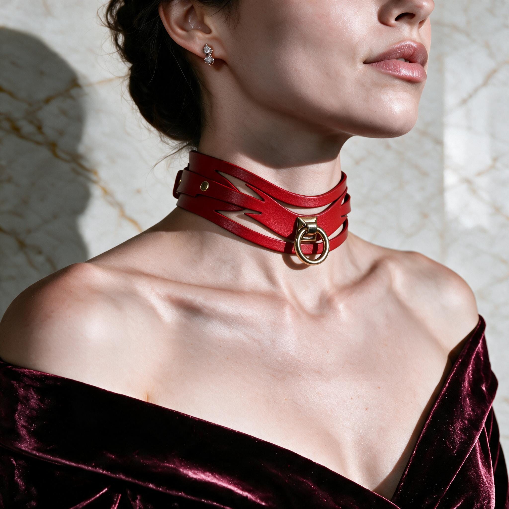 A woman wearing a red choker necklace with a gold buckle, posing against a marble wall.