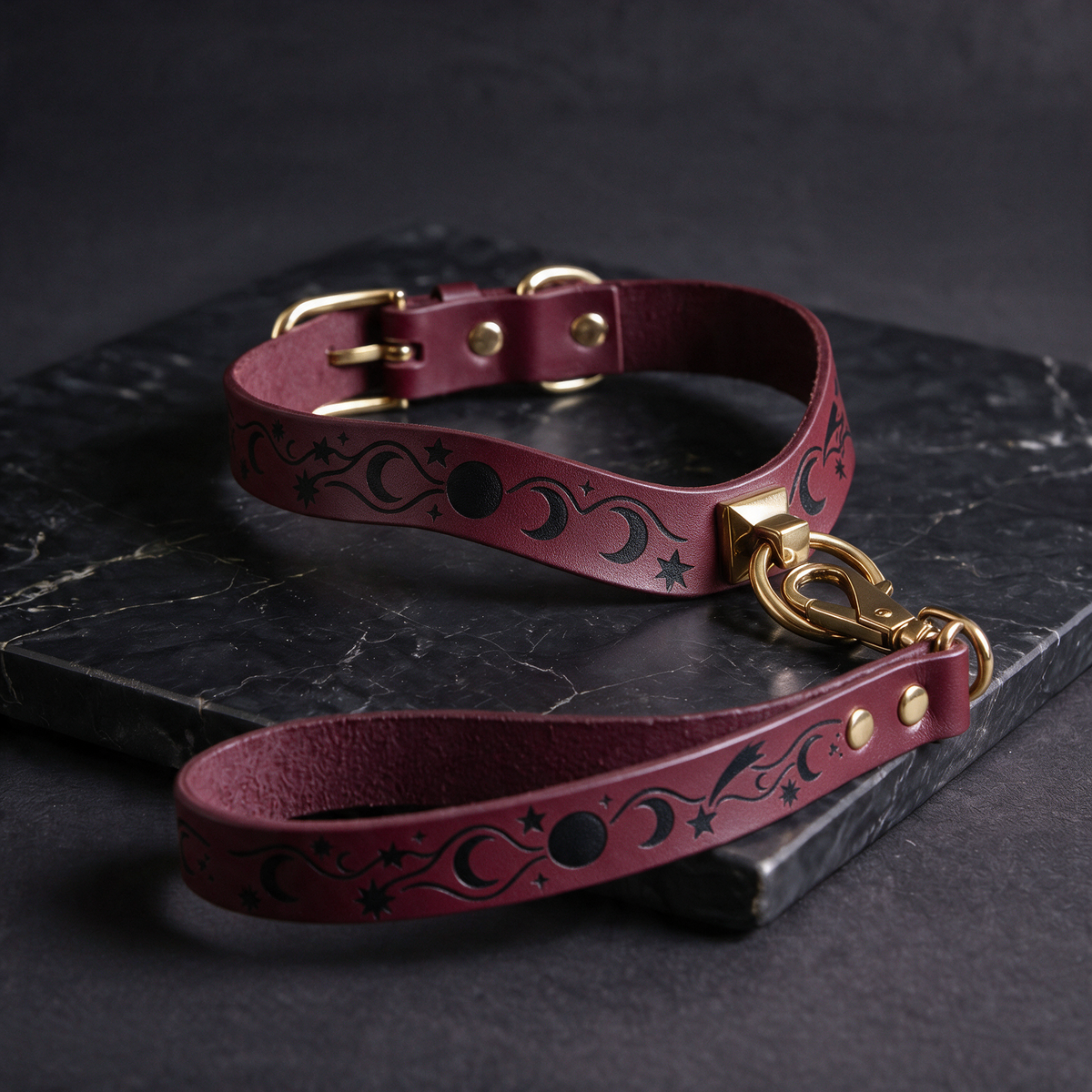 A burgundy leather dog collar with a star and moon design, featuring a gold buckle and a matching leash, is displayed on a black marble surface.