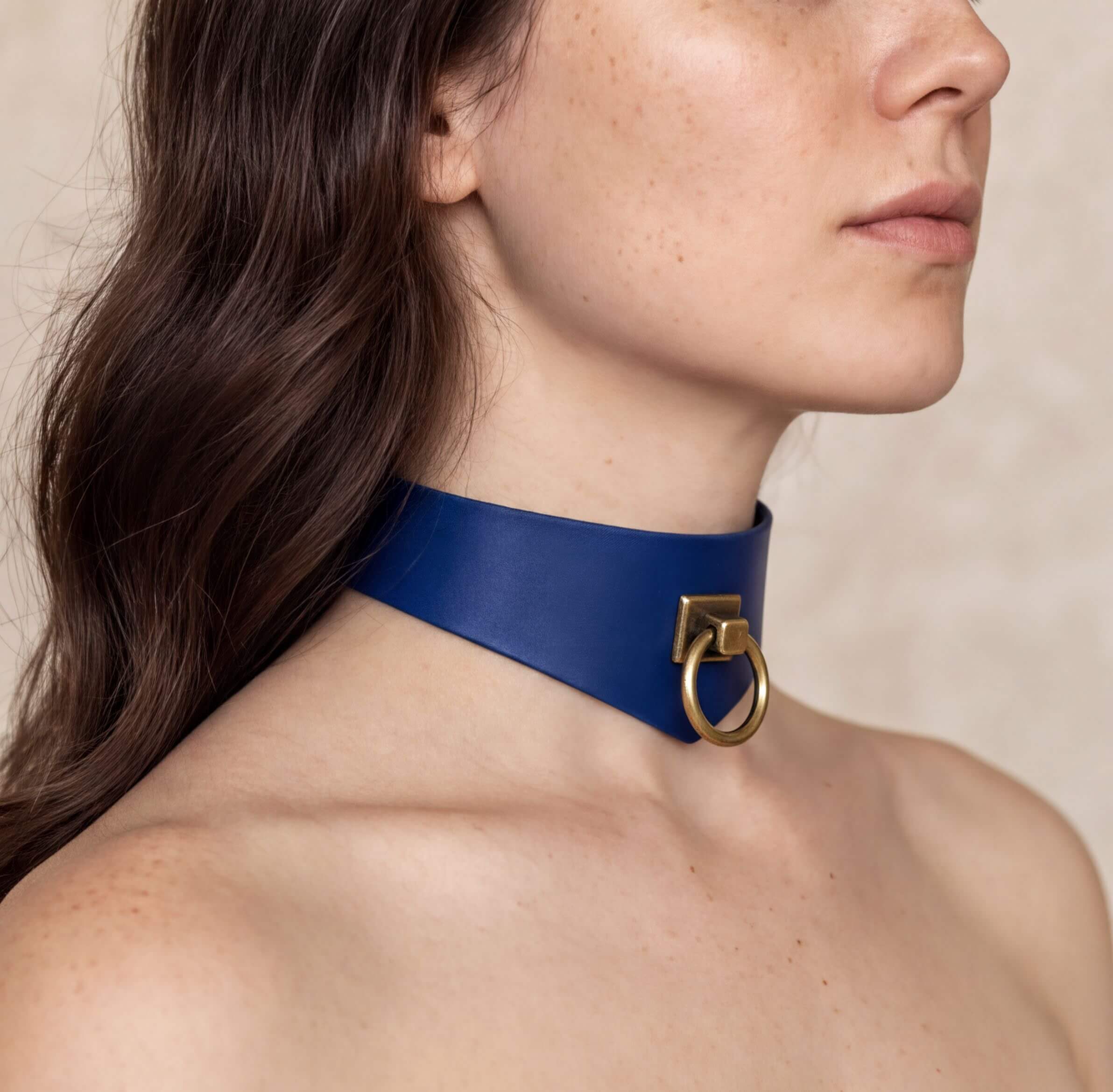 A close-up of a woman's neck and chest area wearing a blue leather choker necklace with a gold-colored metal ring.