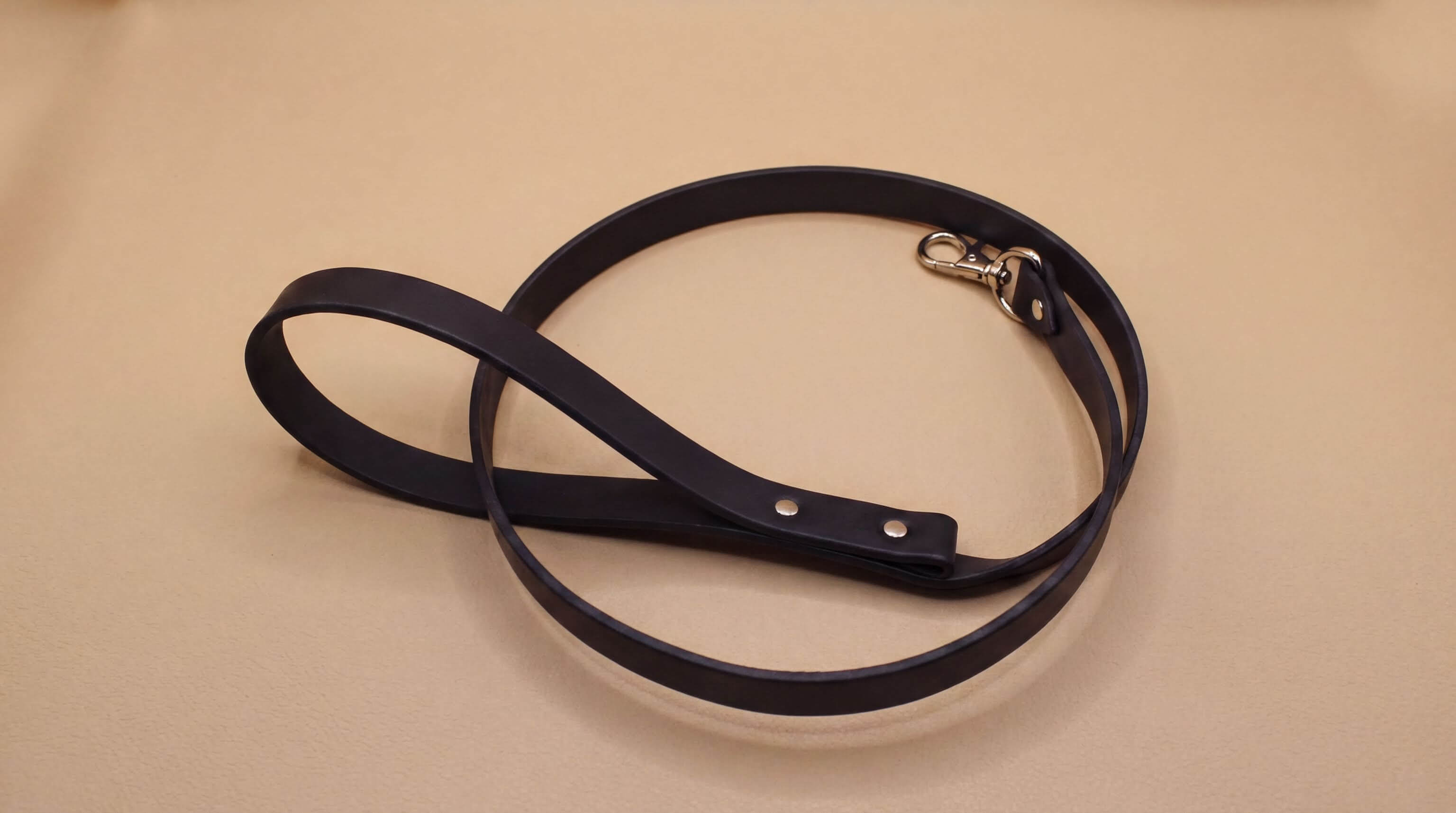 A black leather dog leash with a metal attachment is shown on a beige surface.