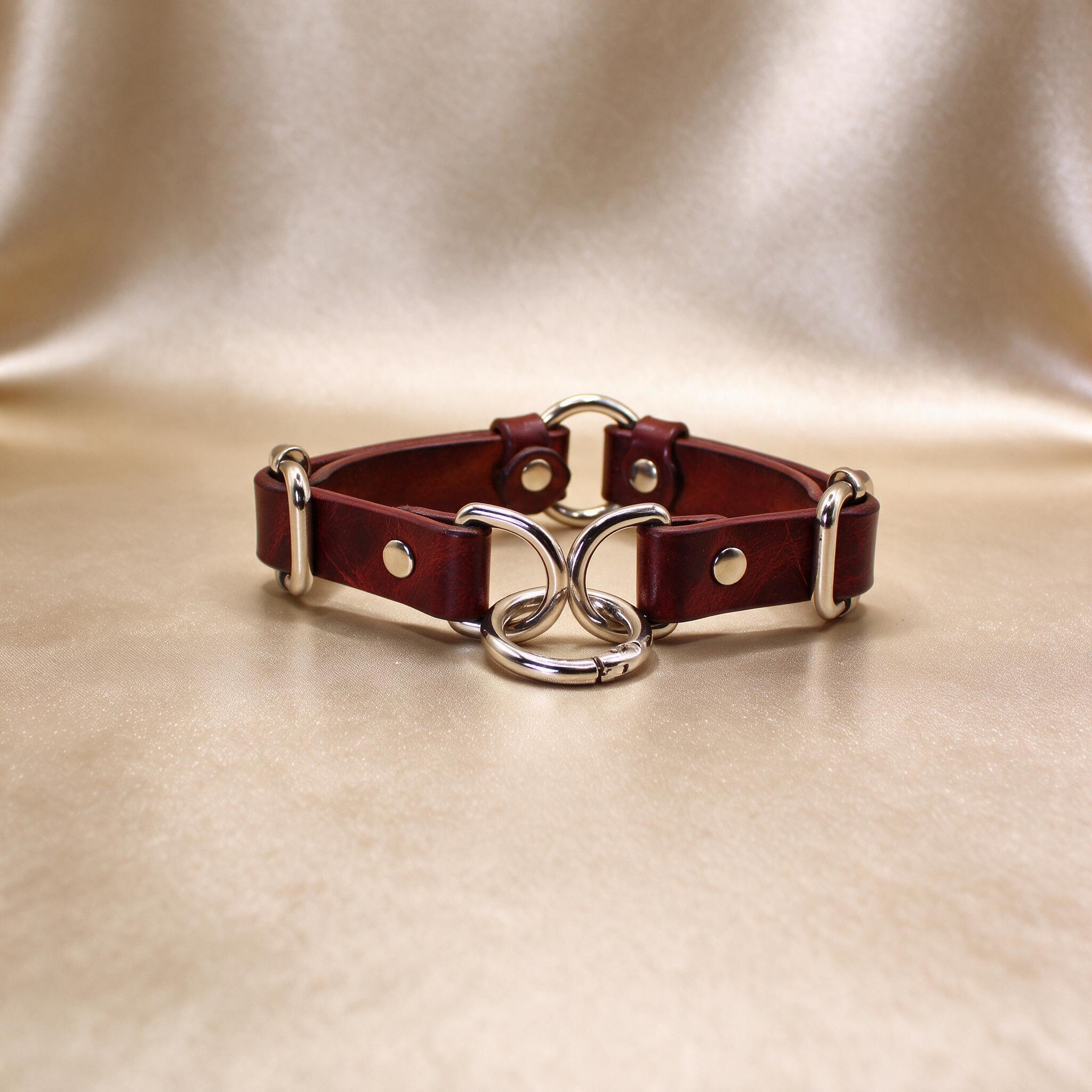 A burgundy leather choker collar with metal rings and buckles, displayed on a beige background.