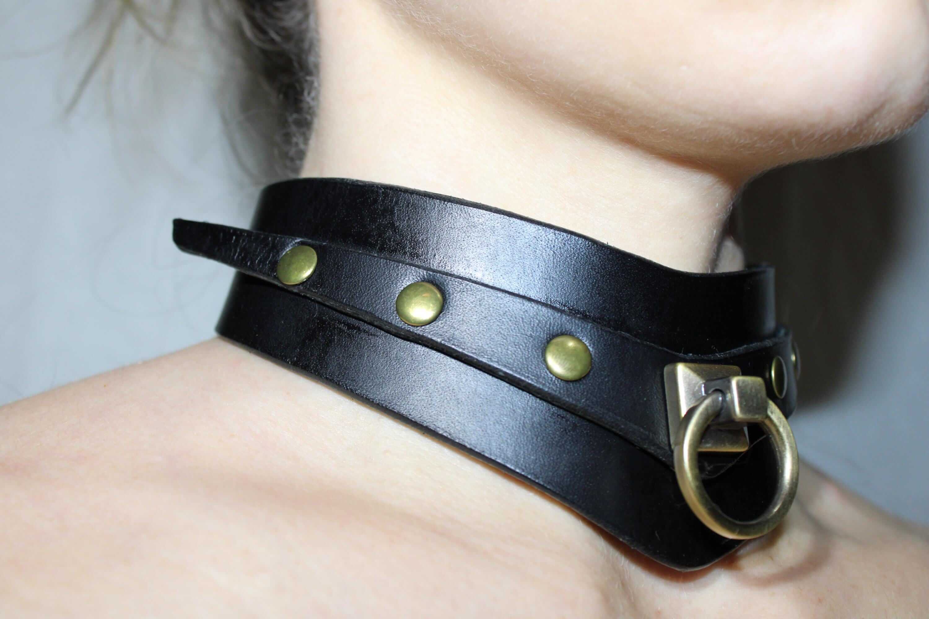 A close-up view of a person's neck wearing a black leather choker necklace with gold-colored studs and a metal ring.