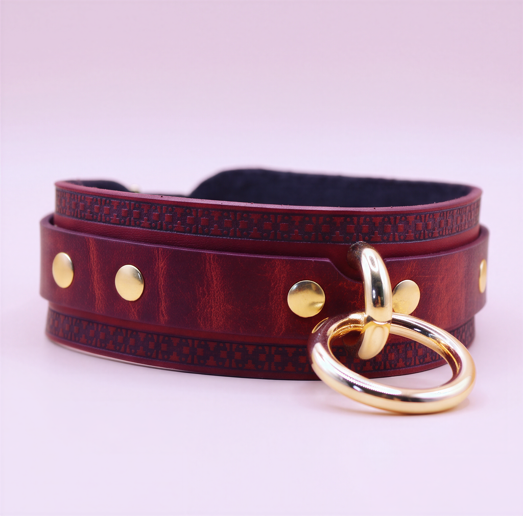 A burgundy leather choker necklace with gold-colored metal studs and a gold-colored metal ring.