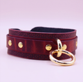 A burgundy leather choker necklace with gold-colored metal studs and a gold-colored metal ring.