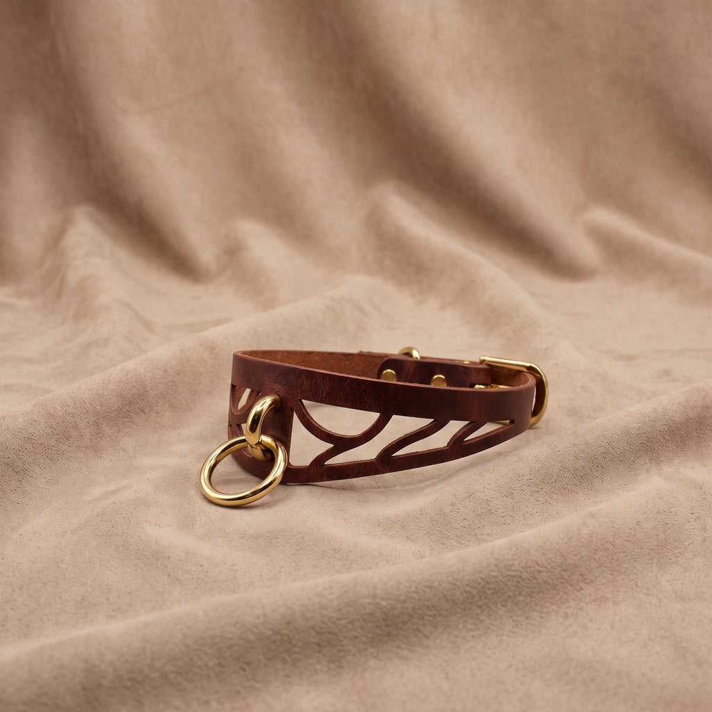 A brown leather choker necklace with a cutout design and two gold-colored rings is displayed on a beige fabric background.