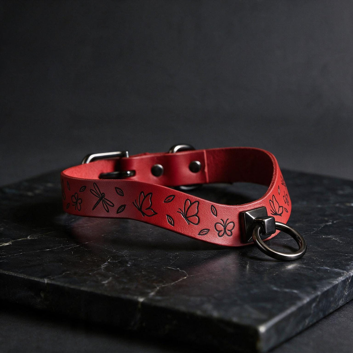 A red leather collar with a metal ring and floral designs on it, resting on a black surface.