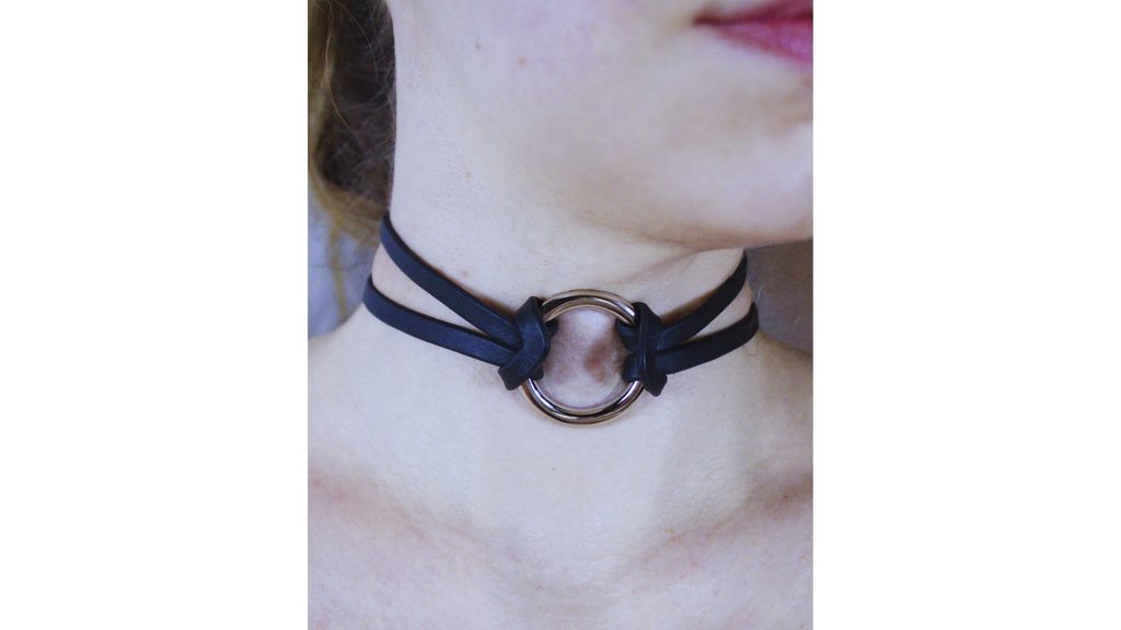 A close-up view of a person's neck and chin area, featuring a black choker necklace with a circular metal ring.