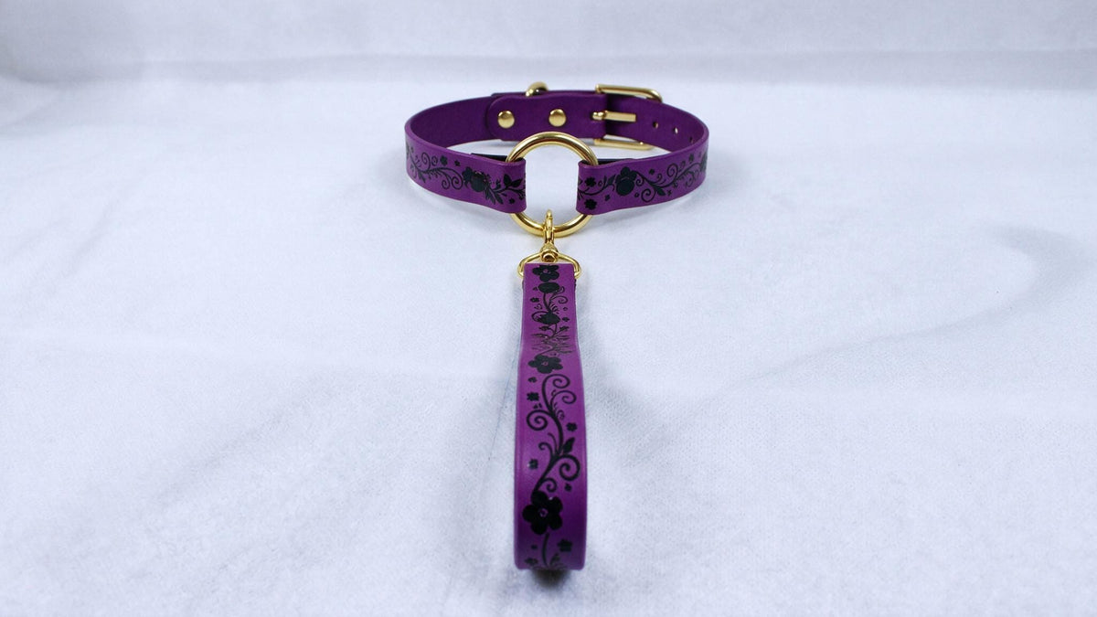 A purple collar with a gold-colored buckle and a floral pattern on the strap is displayed against a white background.