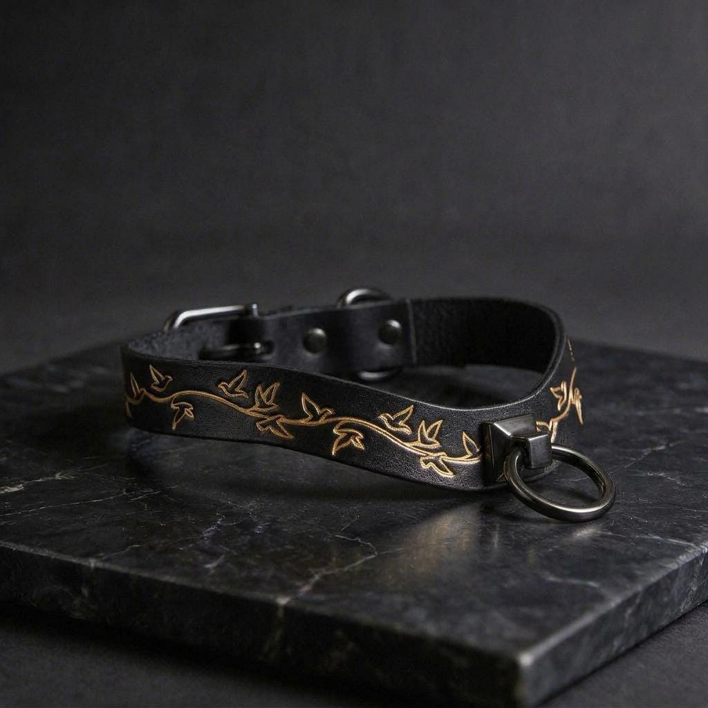 A black leather collar with a gold leaf design, featuring a round metal ring and a metal buckle, is displayed on a black marble surface.