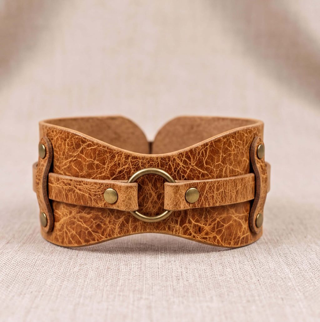 A brown leather corset with a metal ring and two metal fasteners, displayed on a beige fabric background.