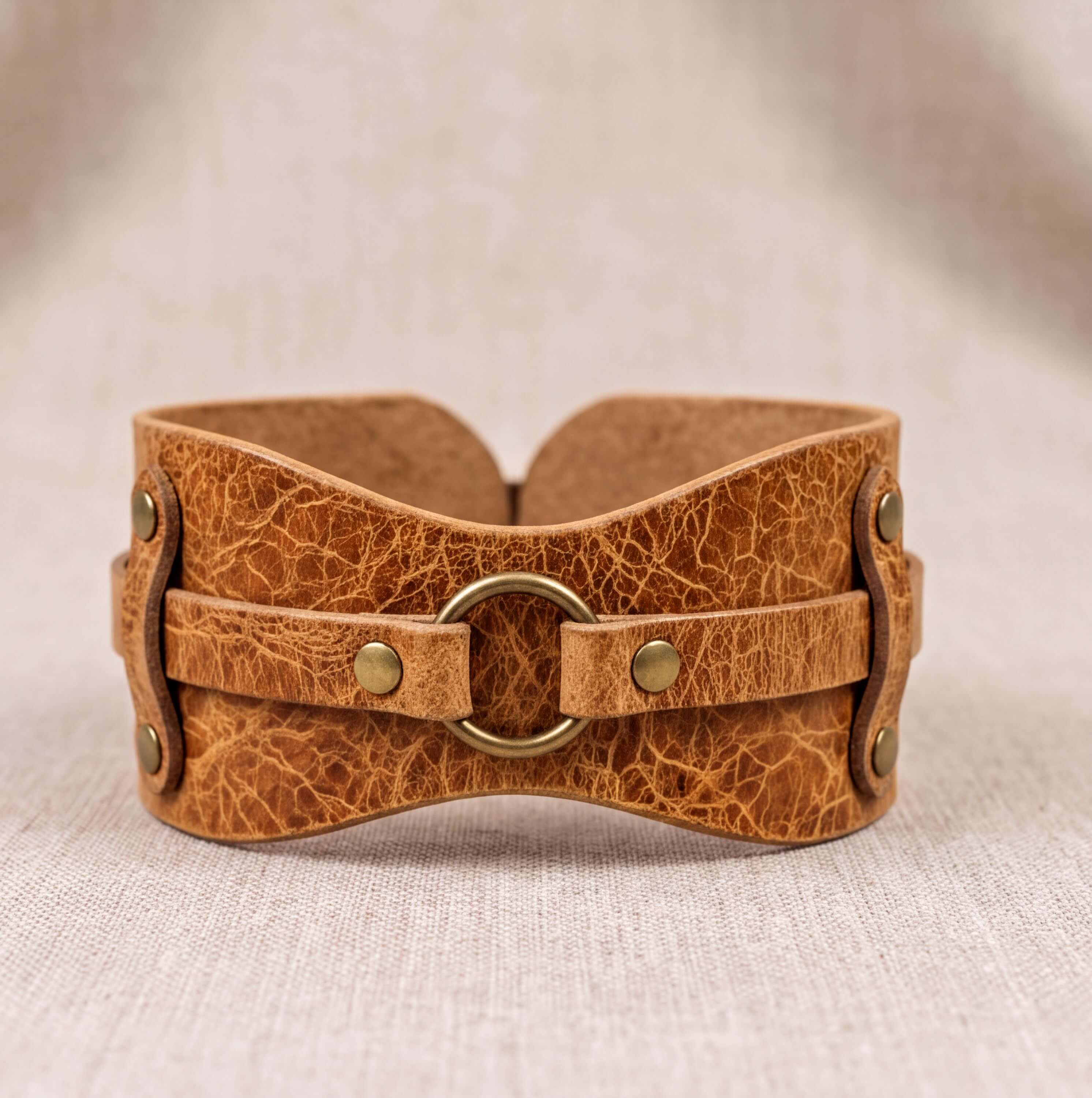 A brown leather corset with a metal ring and two metal fasteners, displayed on a beige fabric background.